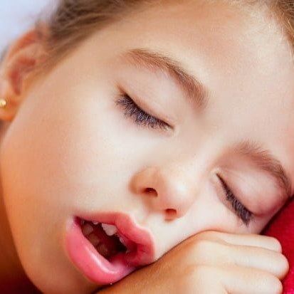 A child with closed eyes and mouth slightly open, sleeping peacefully with their cheek resting on their hand.