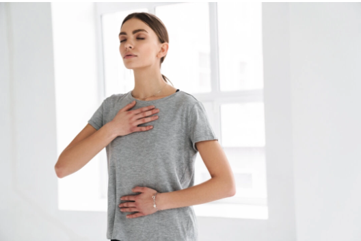 A person in a gray t-shirt stands with eyes closed, hands placed on their chest and abdomen, practicing deep breathing.