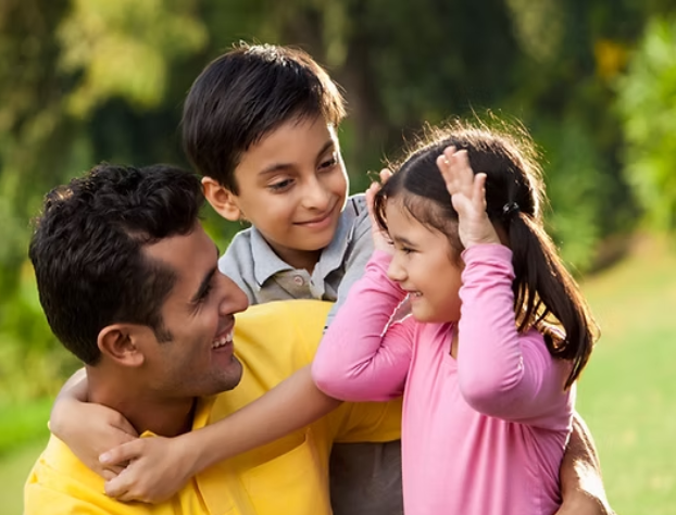 A man in a yellow shirt hugs two children in a park; the girl makes playful bunny ears with her hands.
