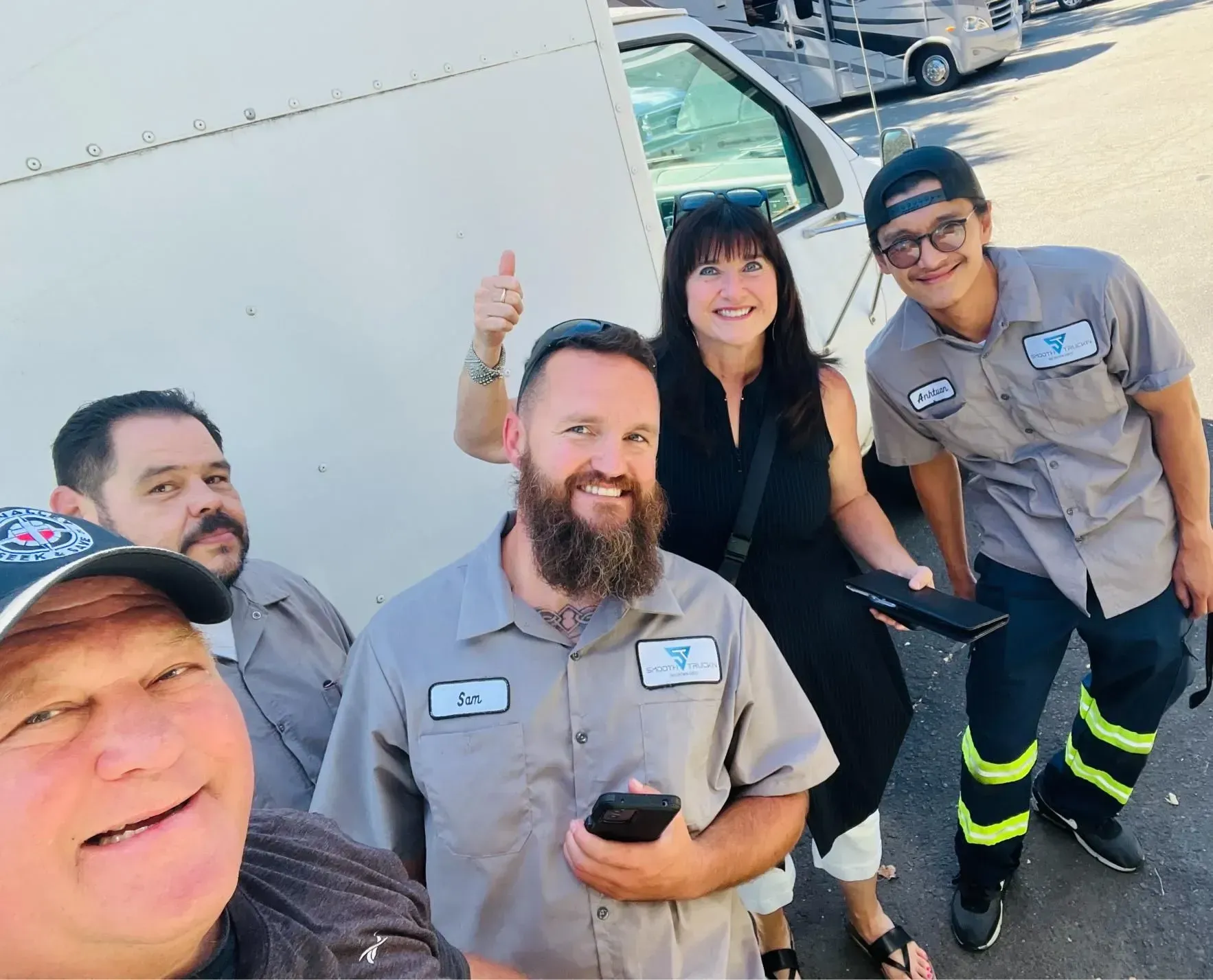 Six people in gray work uniforms smiling and posing outdoors near a white vehicle.