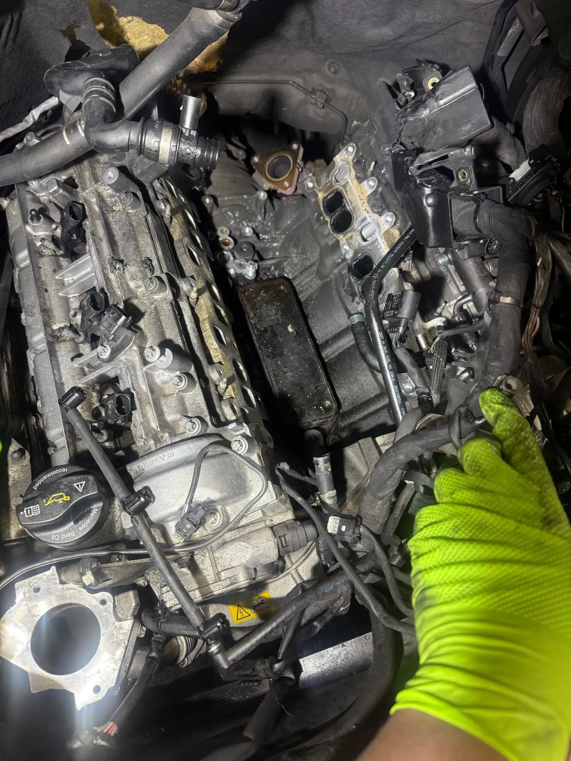 Damaged engine compartment with broken black parts and a gloved hand inspecting debris