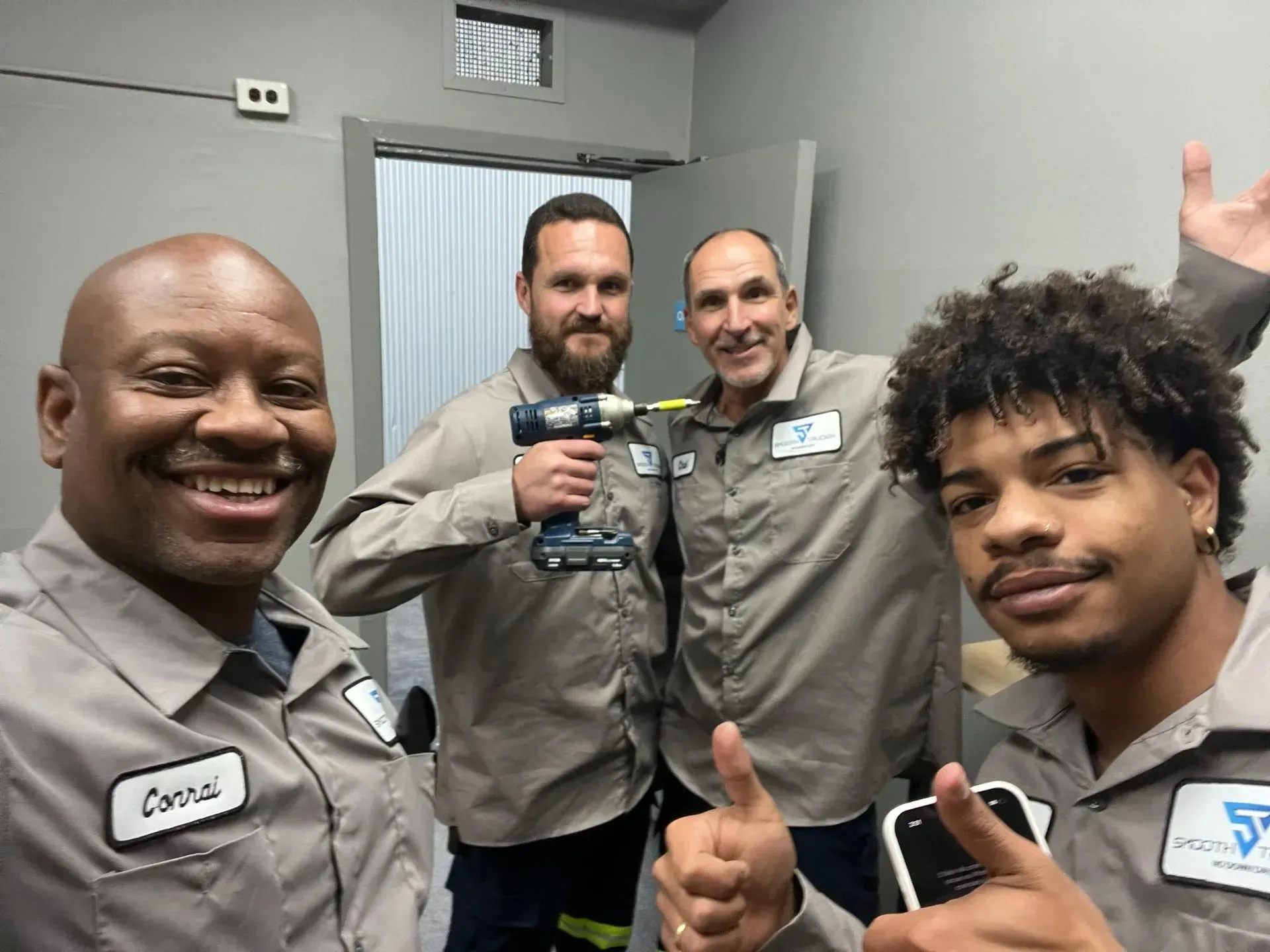 Four coworkers in gray uniforms smile and give thumbs up in a small office room.