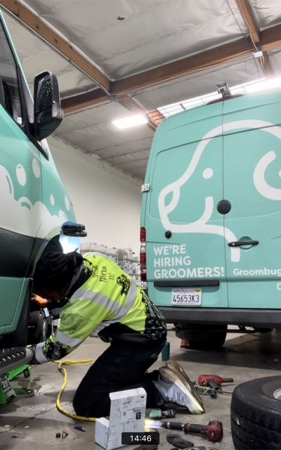 Person kneeling under a turquoise Groombug fleet van, working on the front tire. Wearing neon yellow vest, in garage.