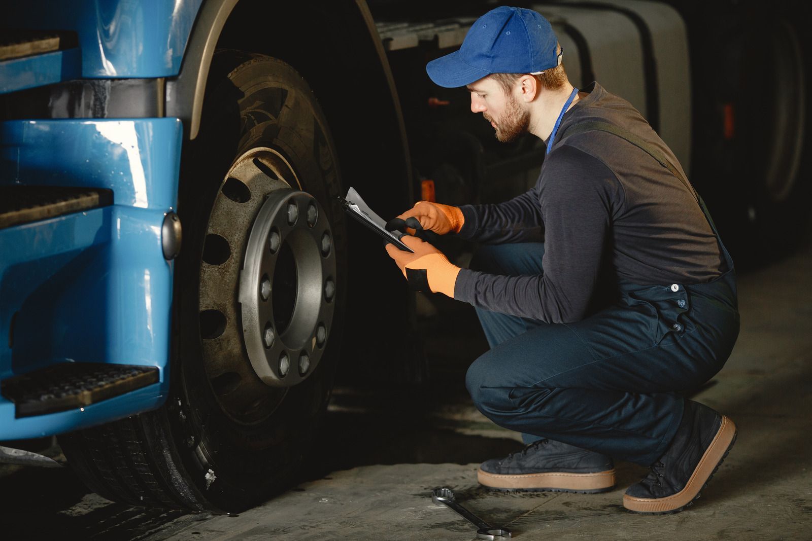 Mechanic in blue cap checks a truck tire with a tool in a dim garage