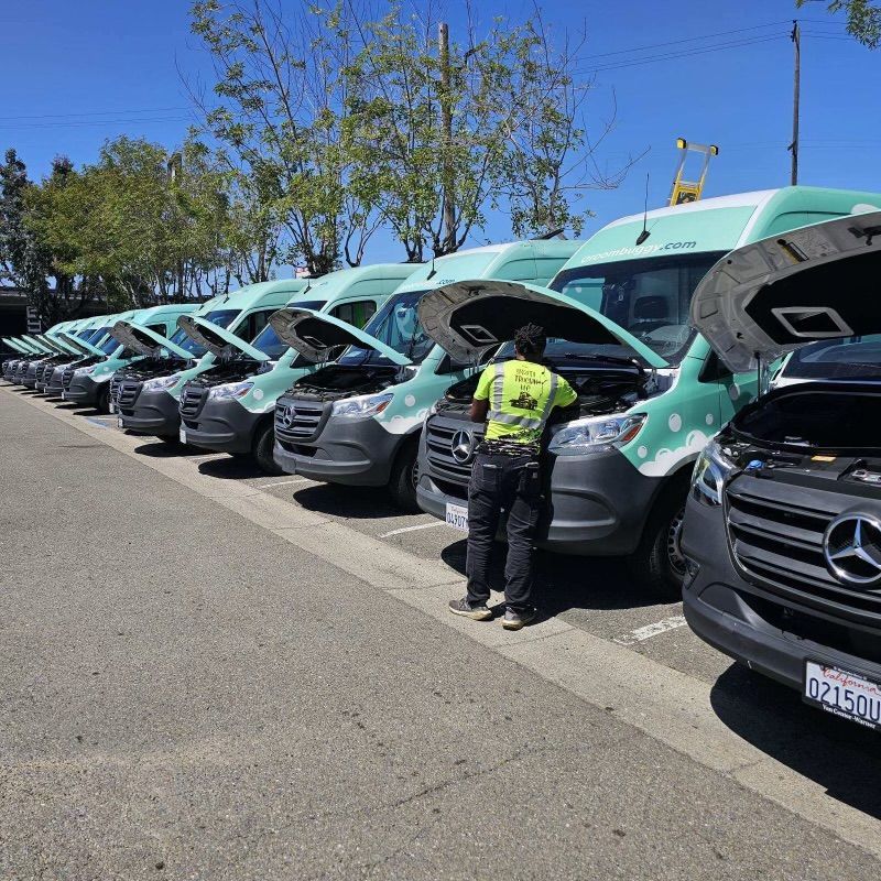 A person in a yellow vest inspects a line of vans with open hoods in a parking lot.