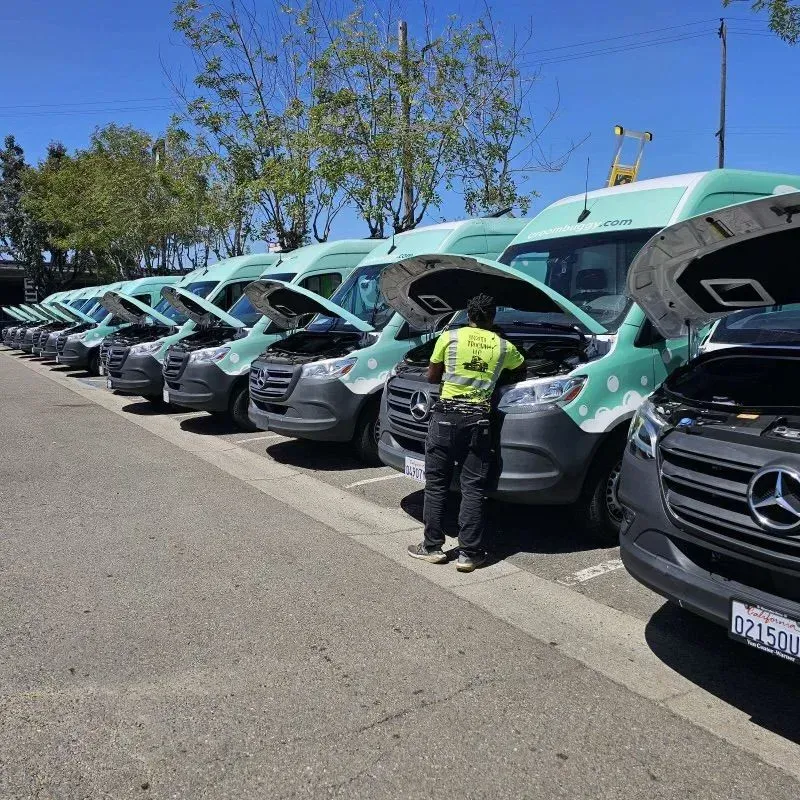 Row of parked Mercedes vans with hoods open while a worker in a safety vest inspects them outside.