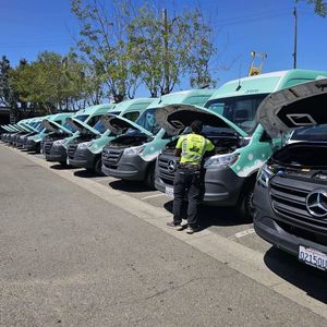 A line of green and silver fleet vans with open hoods. A worker in a yellow vest is inspecting one. Sunny parking lot.