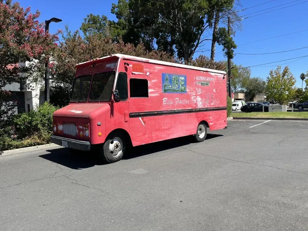 Red food truck parked on a sunny street beside trees and buildings.