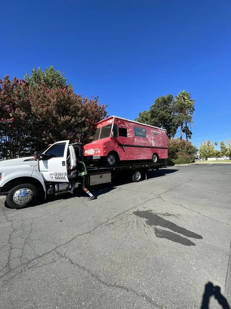 White tow truck loading a red box truck on a flatbed in a parking lot under a clear blue sky