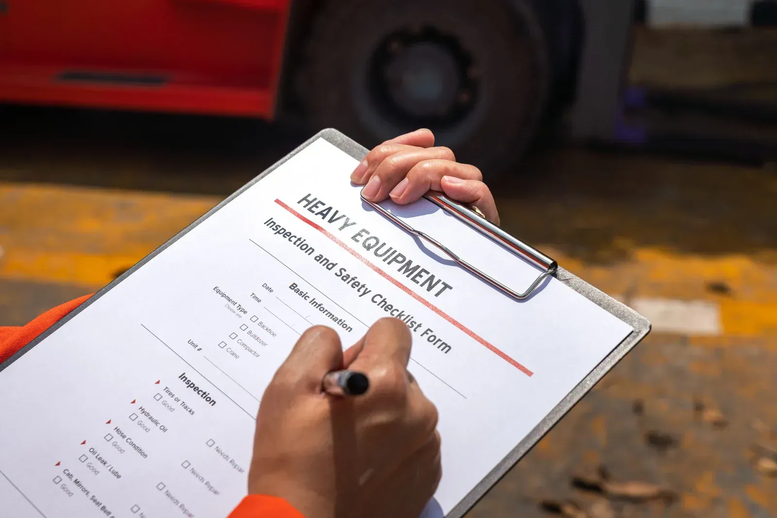 Hand filling out a heavy equipment inspection form on a clipboard beside a truck wheel