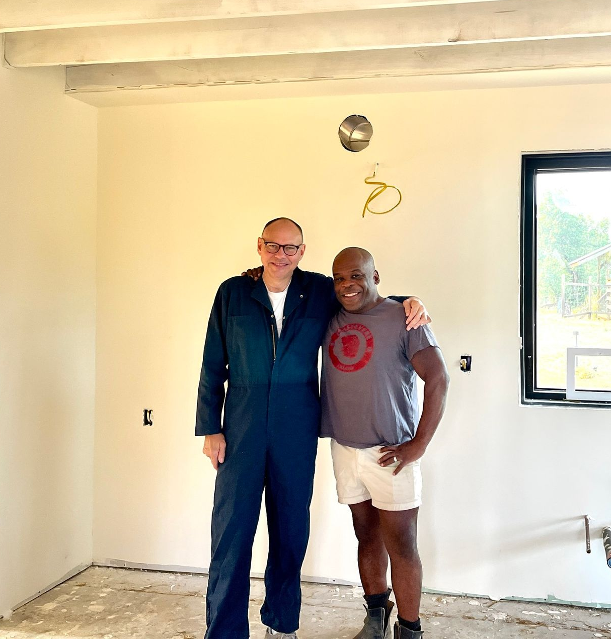 Two people in a room under construction, one wearing a navy jumpsuit and the other in a gray t-shirt and white shorts.