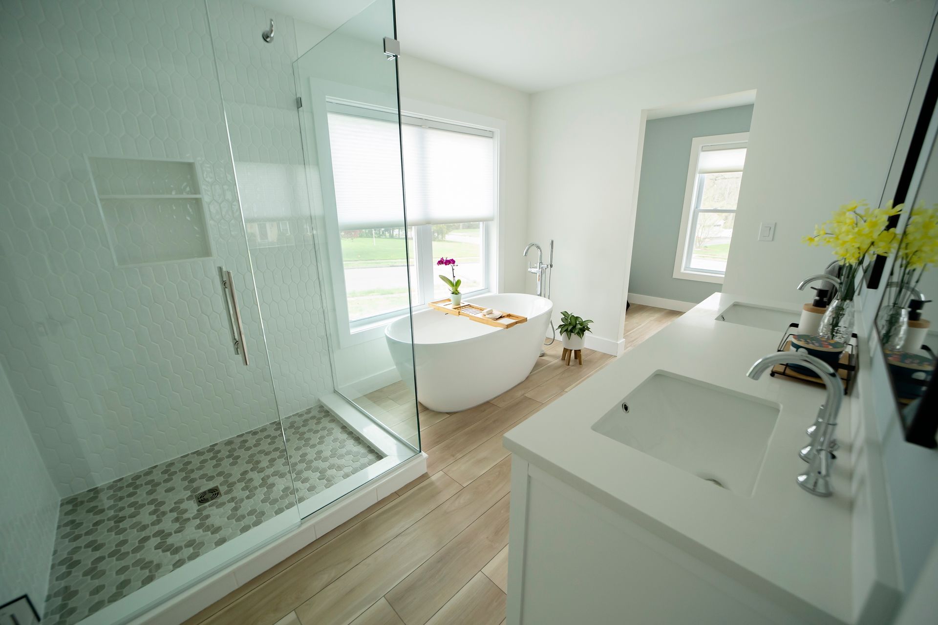 A modern, bright bathroom with a glass-enclosed shower, a white freestanding tub, and a double vanity with stone tops.