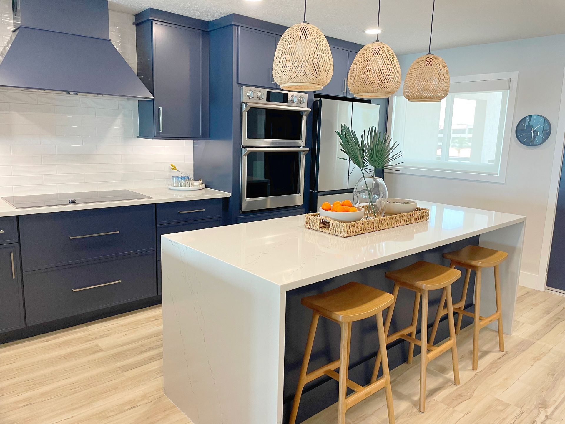 A modern kitchen features navy blue cabinets, a white quartz island with three wooden stools, and woven pendant lights.