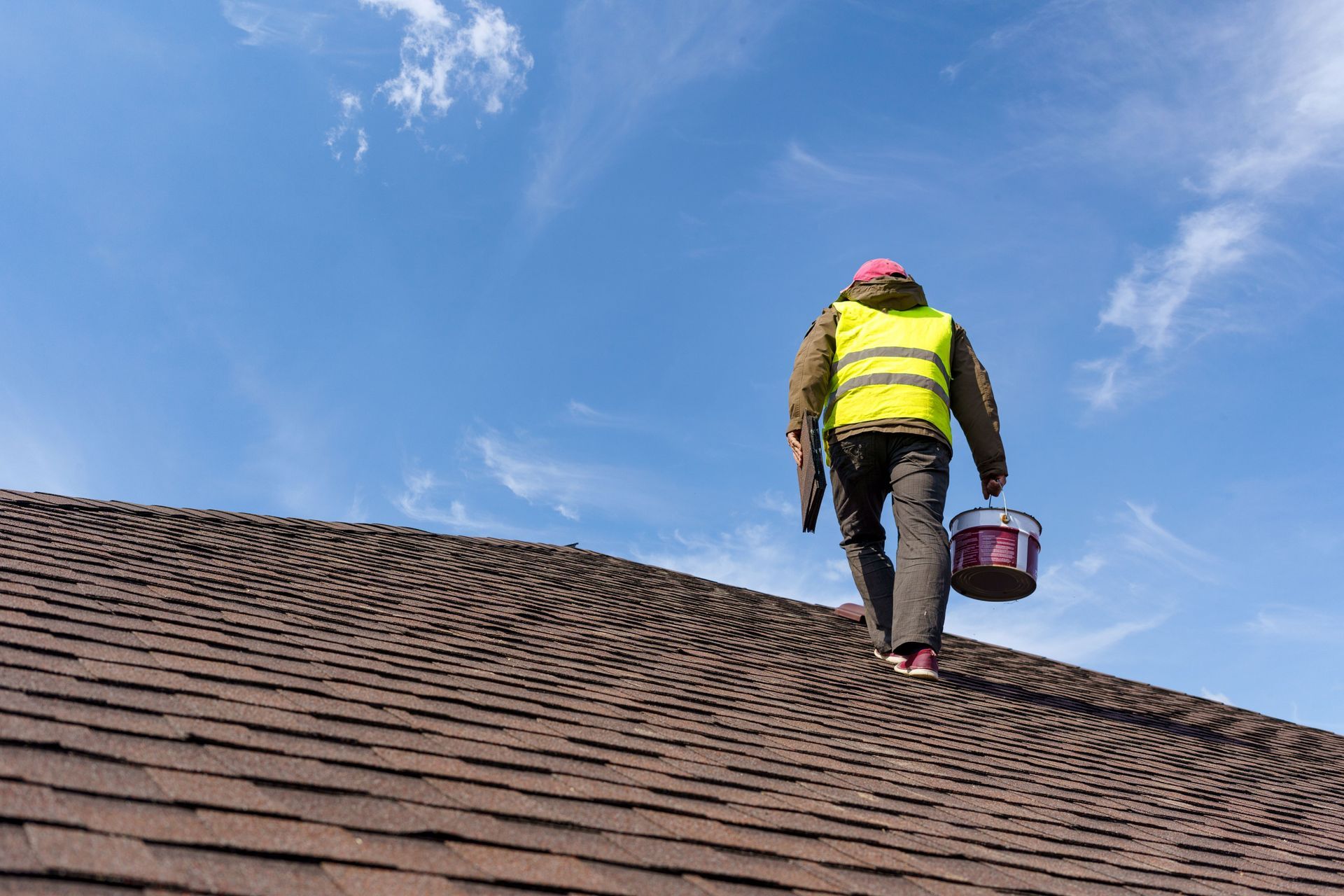 Roofer in safety vest, carrying paint bucket, walks on a shingled roof with a blue sky background.