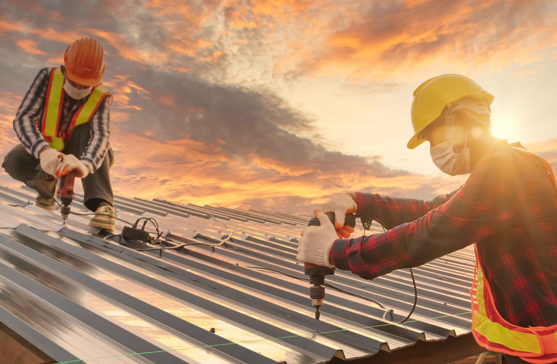 Two construction workers with masks and helmets installing a metal roof at sunset.