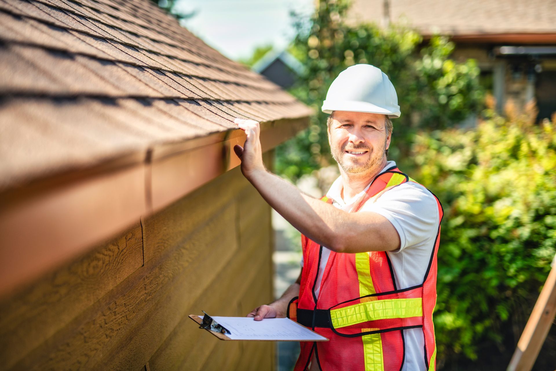 Man in hard hat and safety vest inspecting a building's gutter; he holds a clipboard and smiles.
