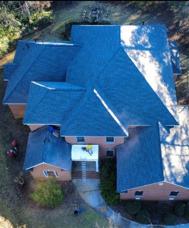 A newly completed residential roofing project by JCB Roofing. Aerial view of a brick house with a dark blue roof, shadowed by trees, and a white porch with a worker.