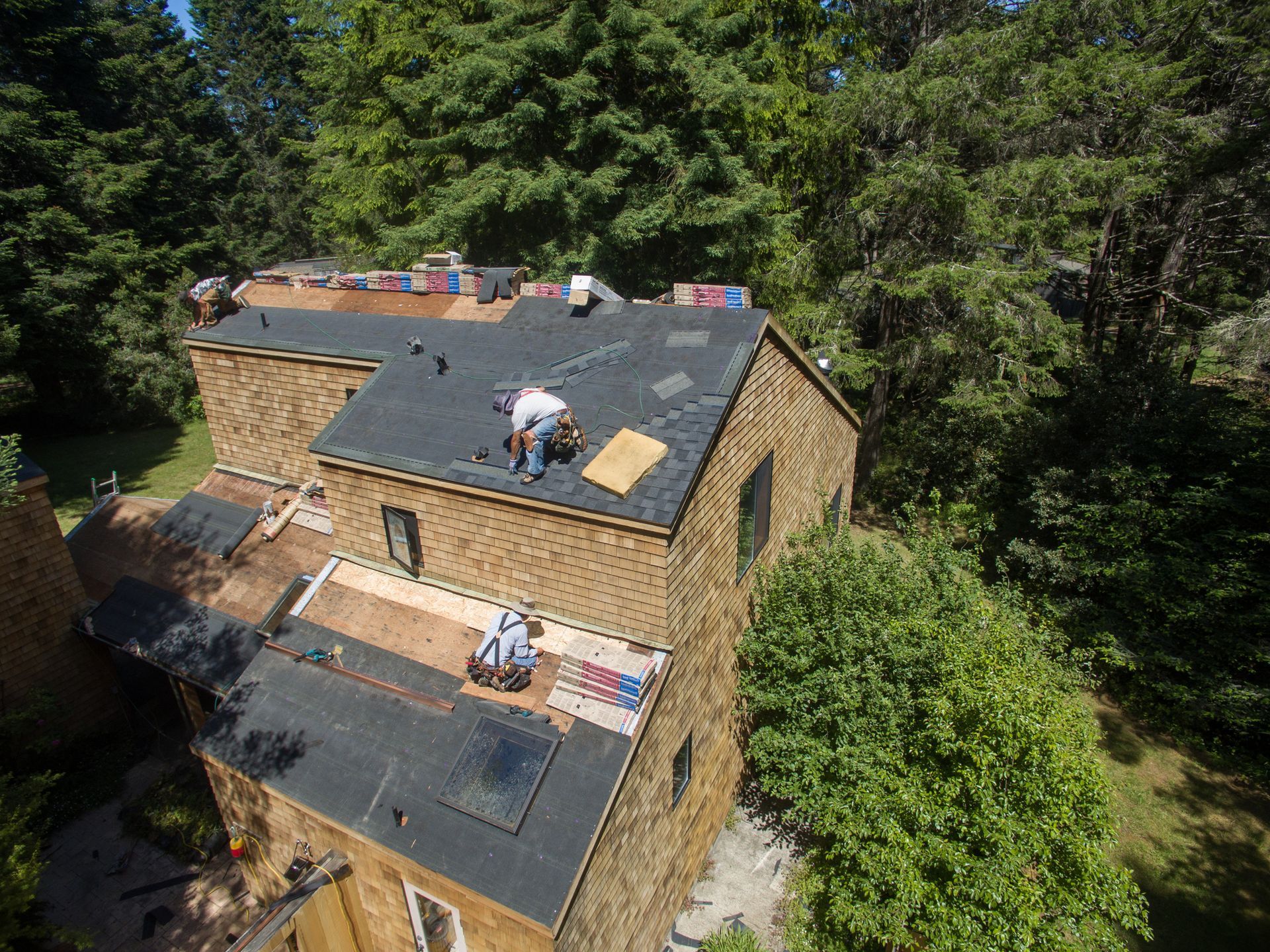 Workers installing shingles on a brick building's roof, surrounded by trees.