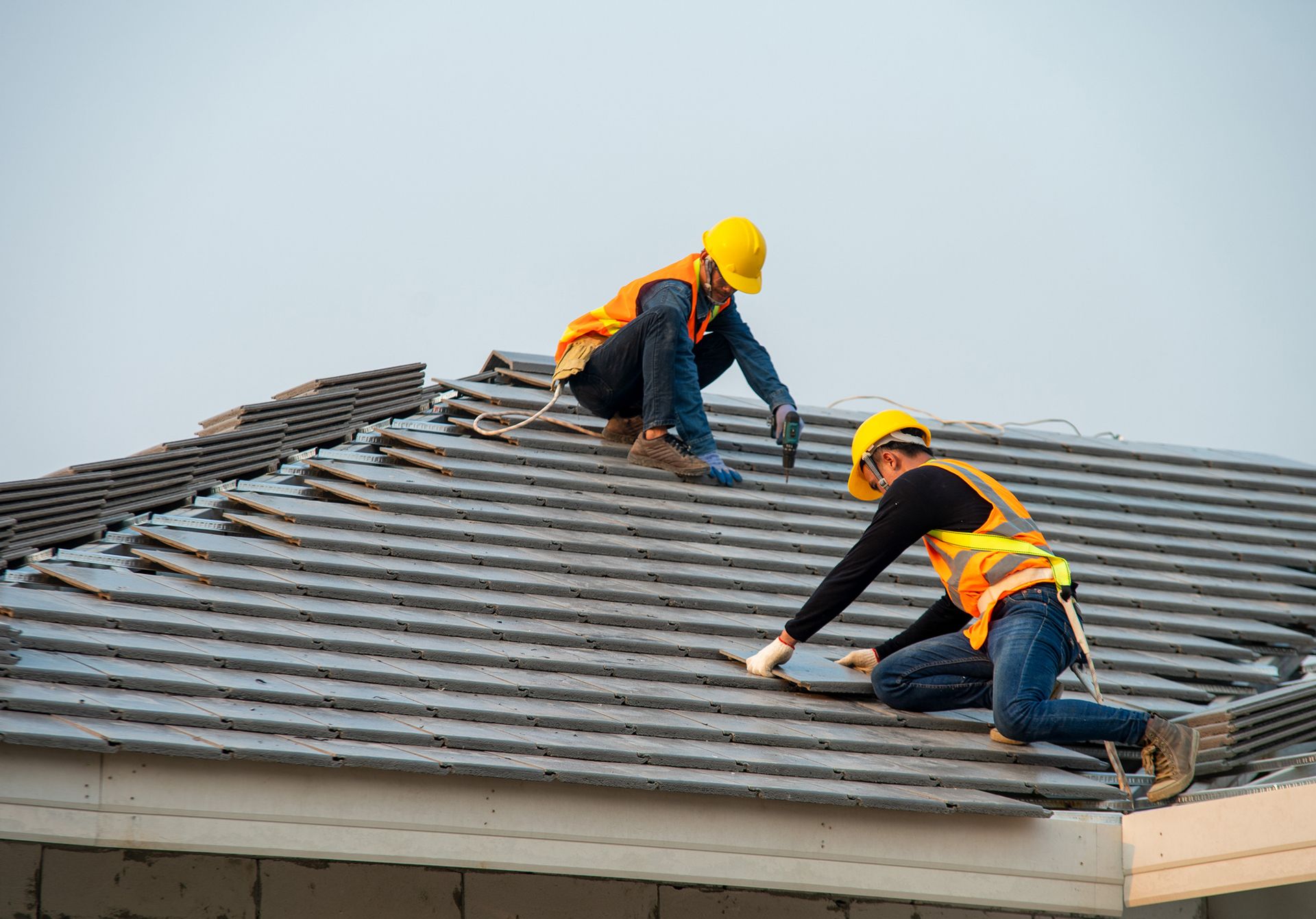 Two roofers inspecting and finishing a new ceramic tile roof installation professionally. Two roofers in hard hats and safety vests installing tiles on a gray roof.