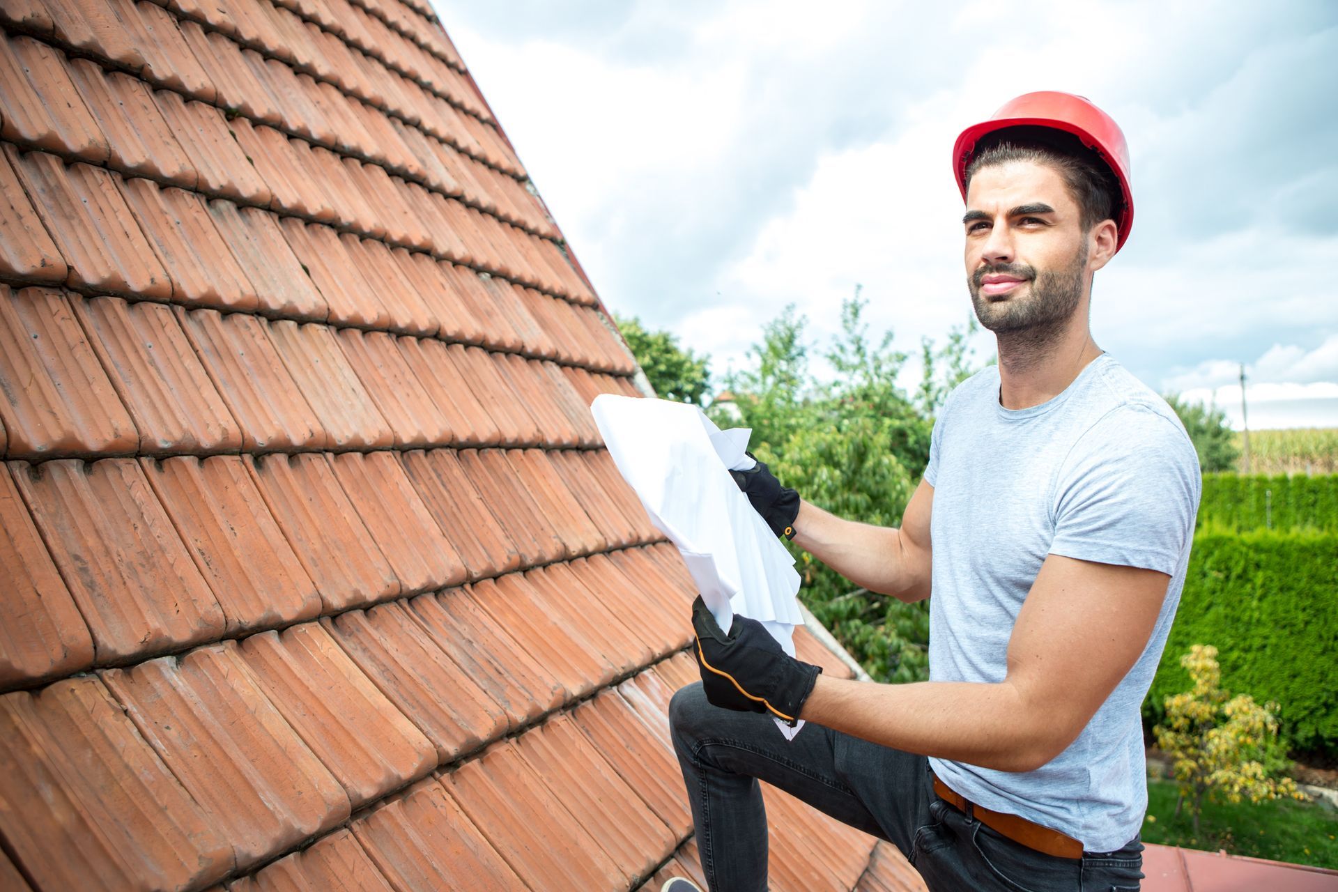 A man is inspecting a roof. Roofer on a tiled roof in a red hard hat, holding a blueprint, smiling. Outdoors.