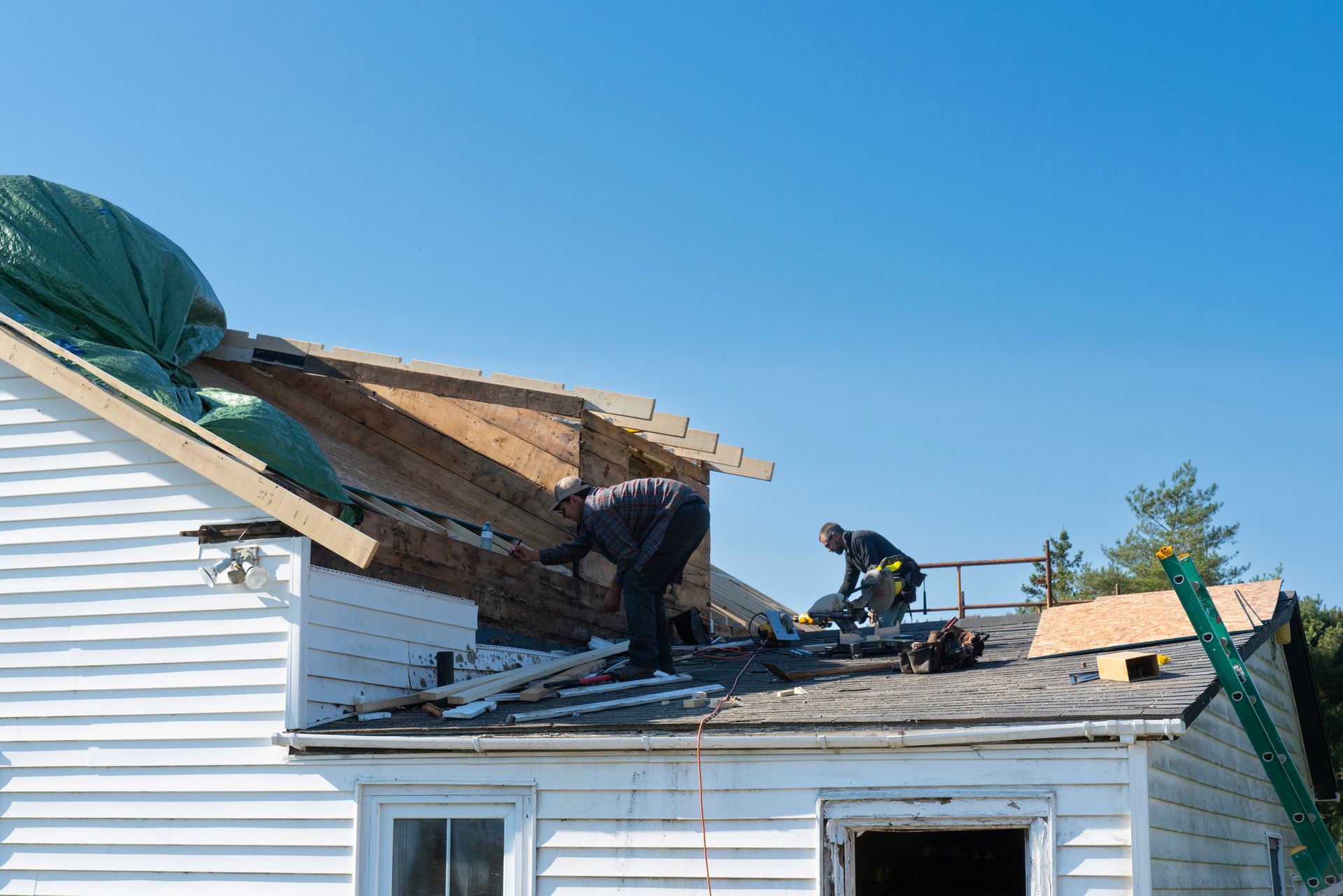 Experienced roofers working on residential roofing repairs. Two workers on a roof, repairing damage on a sunny day. House is white. Blue sky.