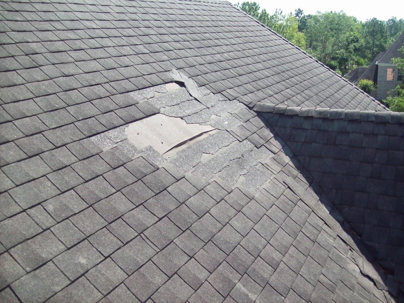 Damaged asphalt shingle roof. Gray shingles show a large area of missing material near a roof intersection.