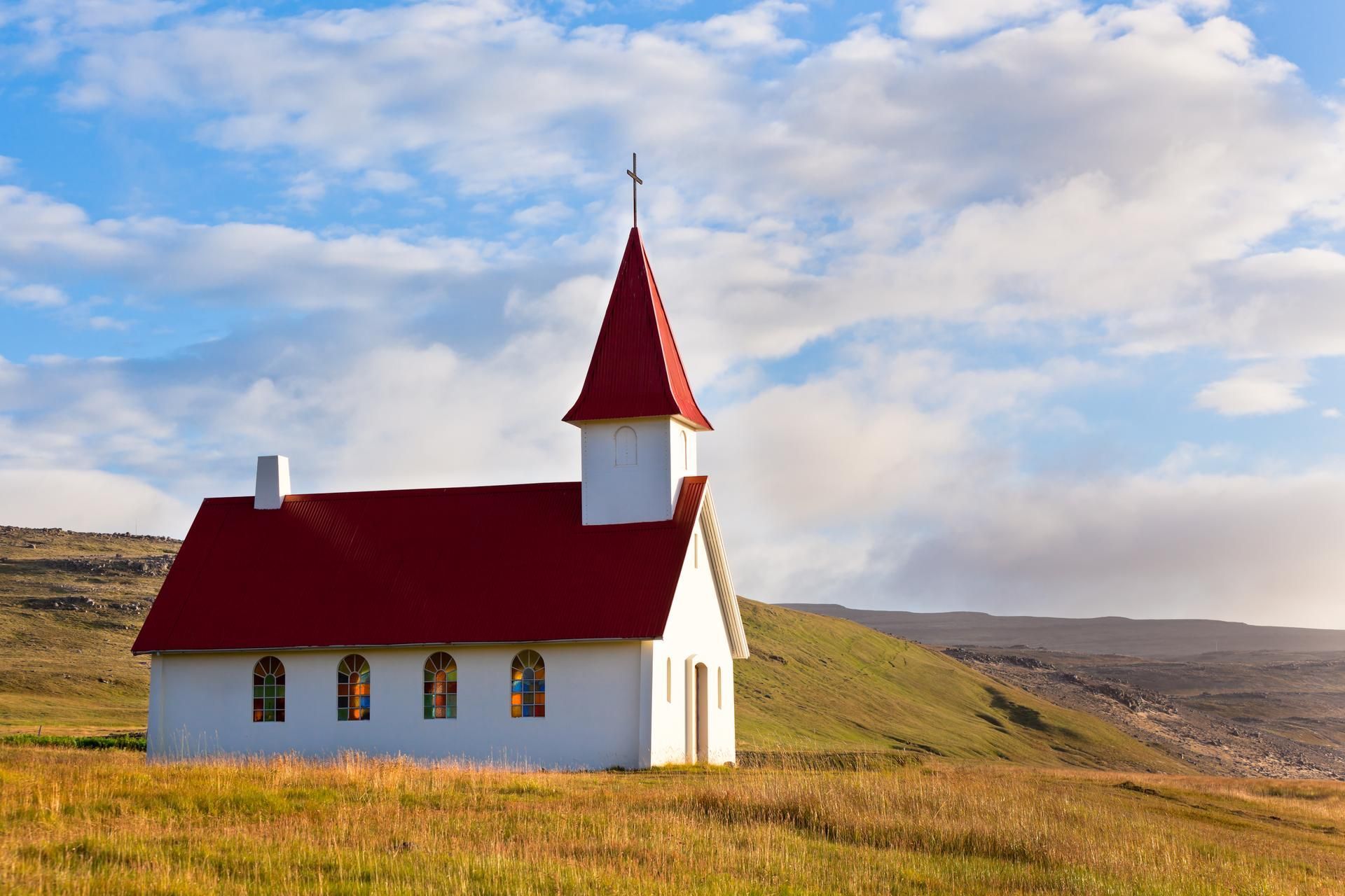 White church with red roof and cross, in a grassy field under a blue sky with clouds.