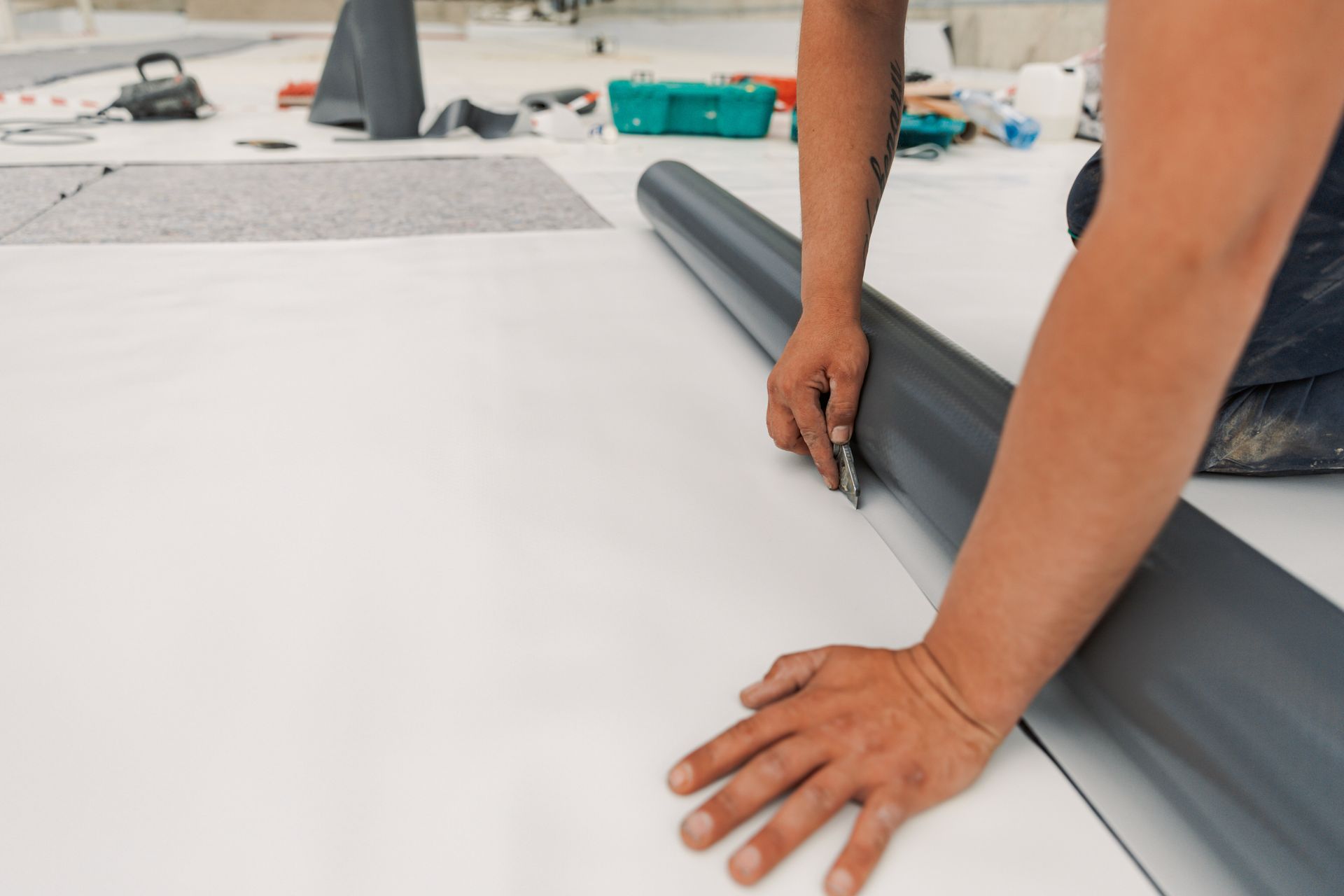 Person cutting rolled flooring on a white surface with tools visible in the background.