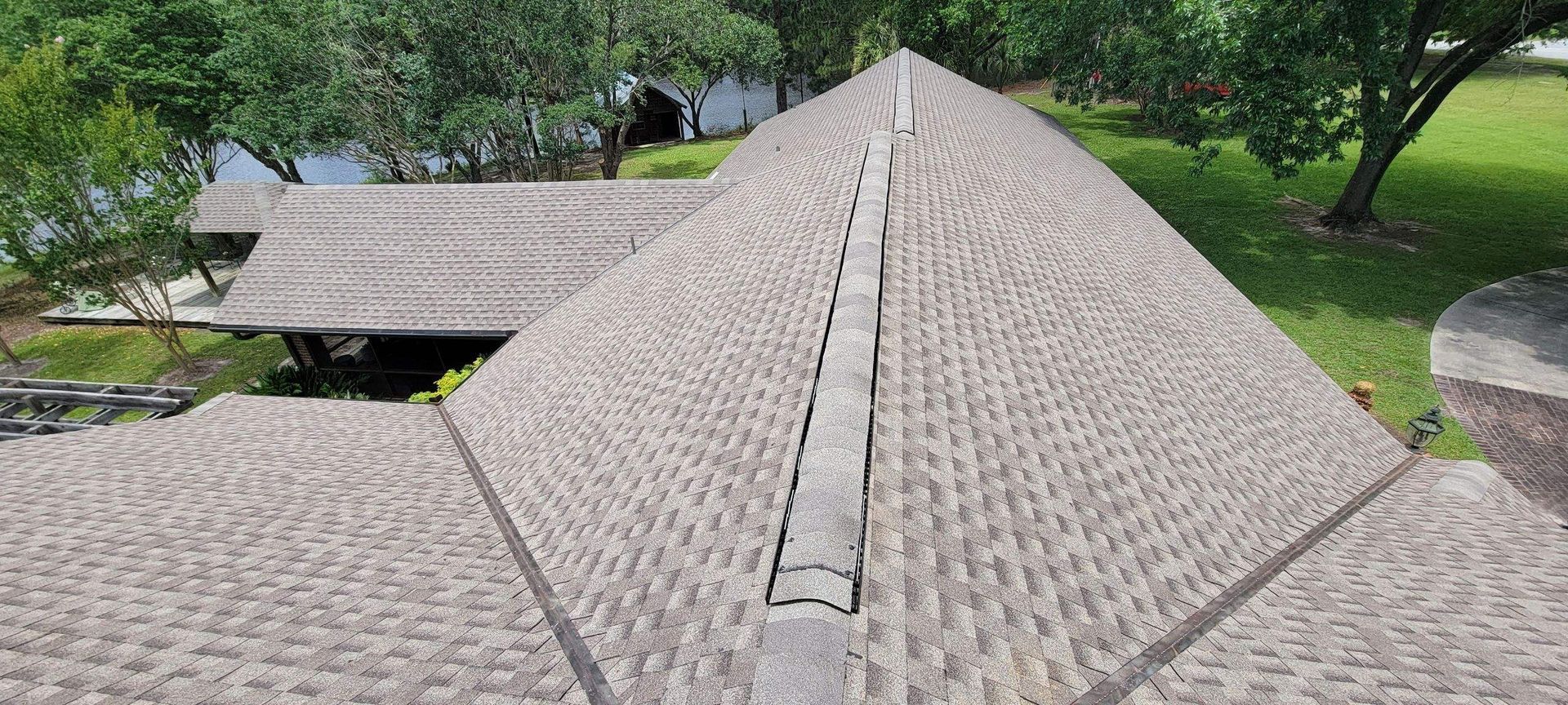 Overhead view of a roof with weathered shingles, centered by a concrete ridge. Green trees and grass are in the background.