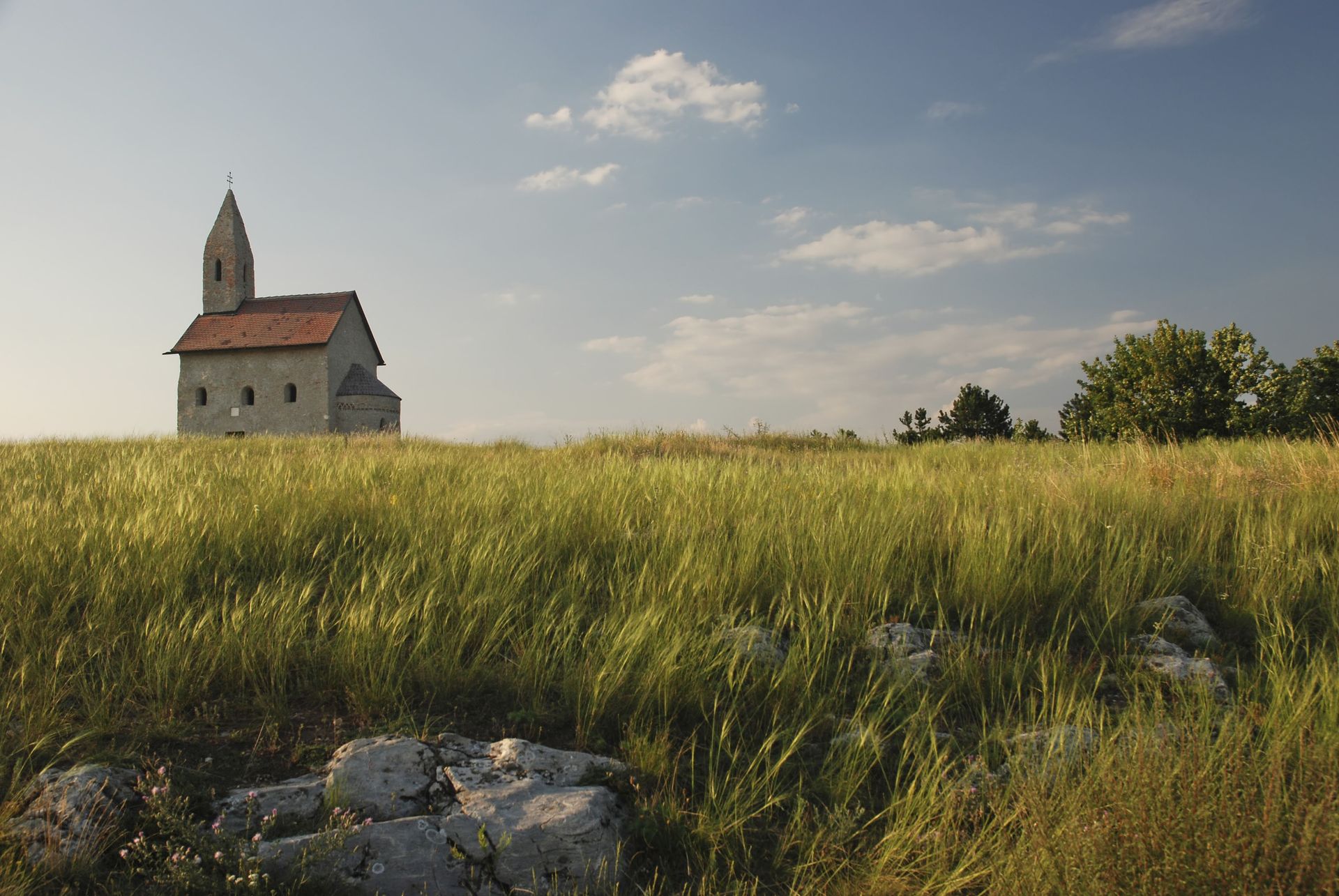 Stone church with a bell tower on a grassy hill, under a blue sky with some clouds.