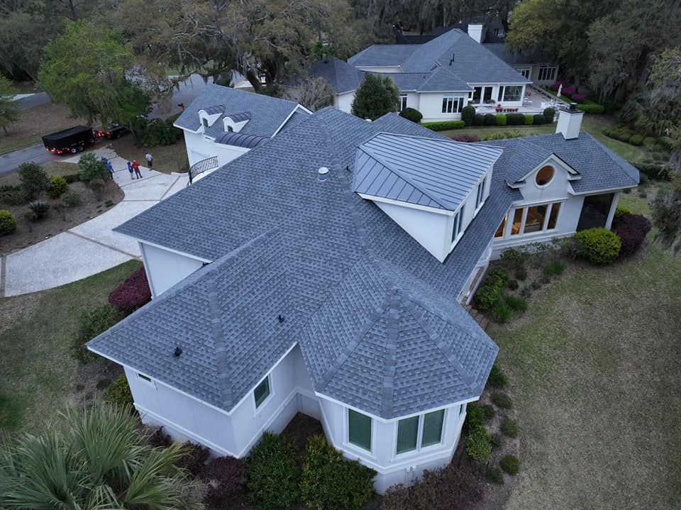 Aerial view of a large white house with a multi-gabled gray shingle roof and a metal-roofed dormer, set in a wooded area.