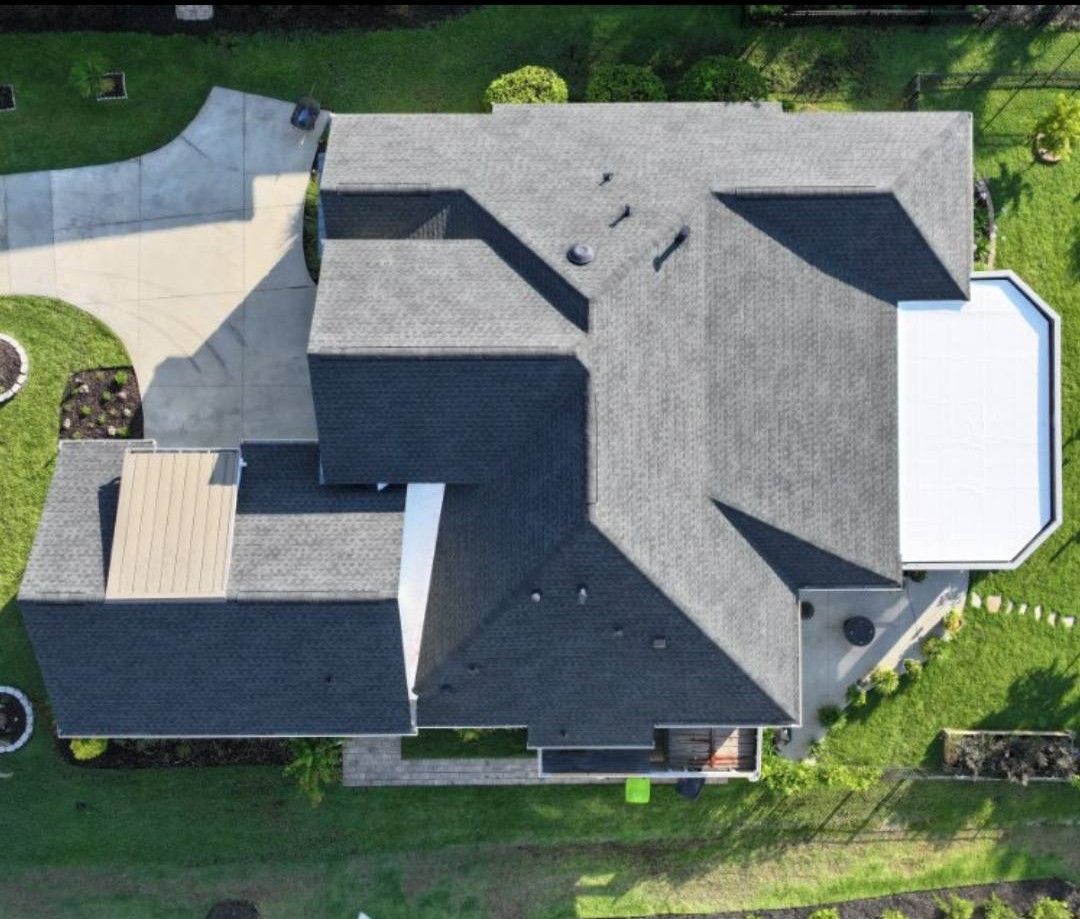 Aerial view of a residential house with a dark gray shingled roof, a white patio cover, and a driveway on a grassy lawn.