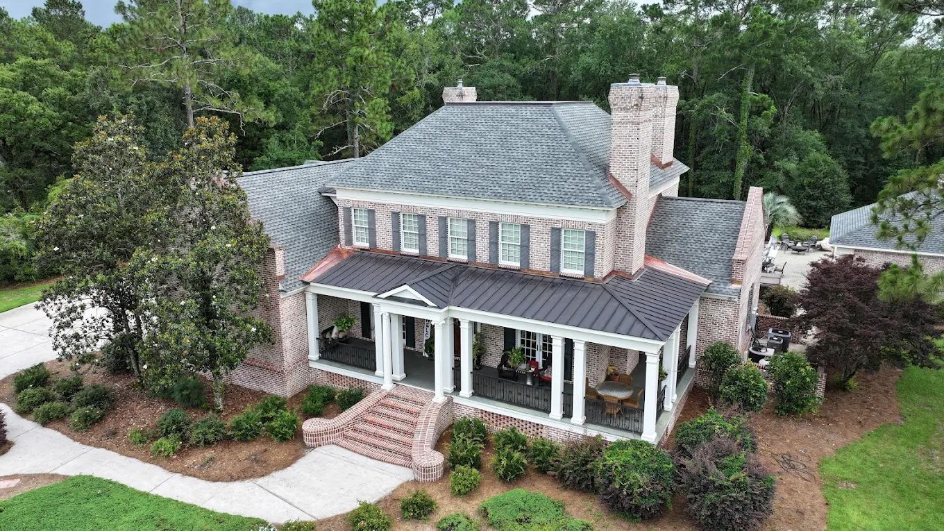 An aerial view of a two-story brick house with a dark metal porch roof, light-colored trim, and surrounding landscaping. An aerial view of a two-story brick house with a dark metal porch roof, light-colored trim, and surrounding landscaping.