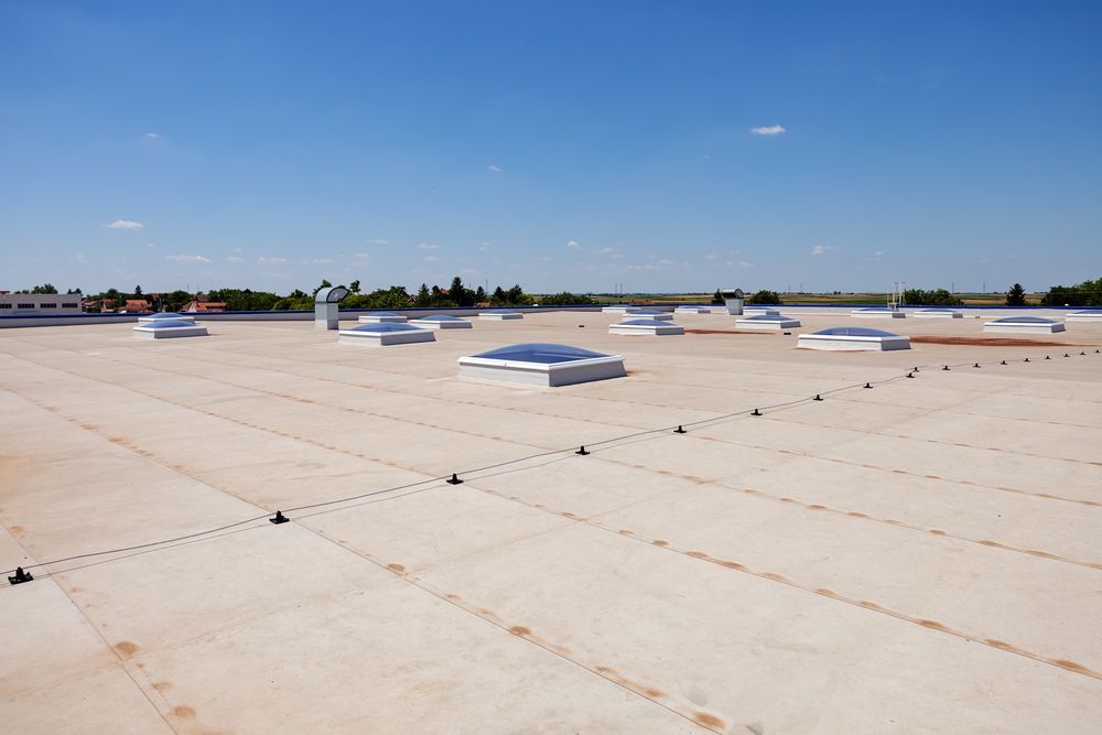 Flat, light-colored commercial roof with several skylights, under a bright blue sky.