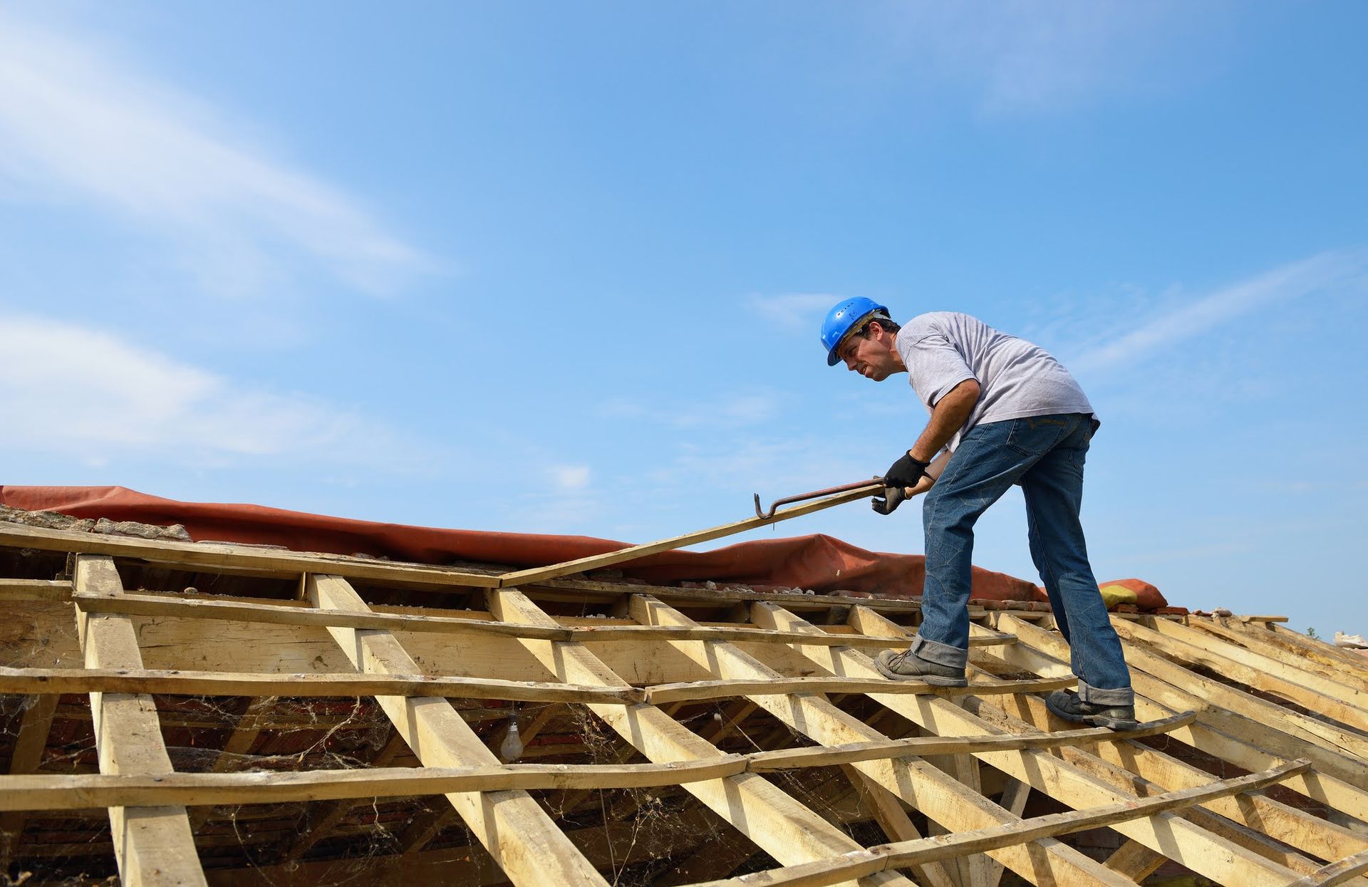 Man in blue hardhat on a roof uses a tool; blue sky.