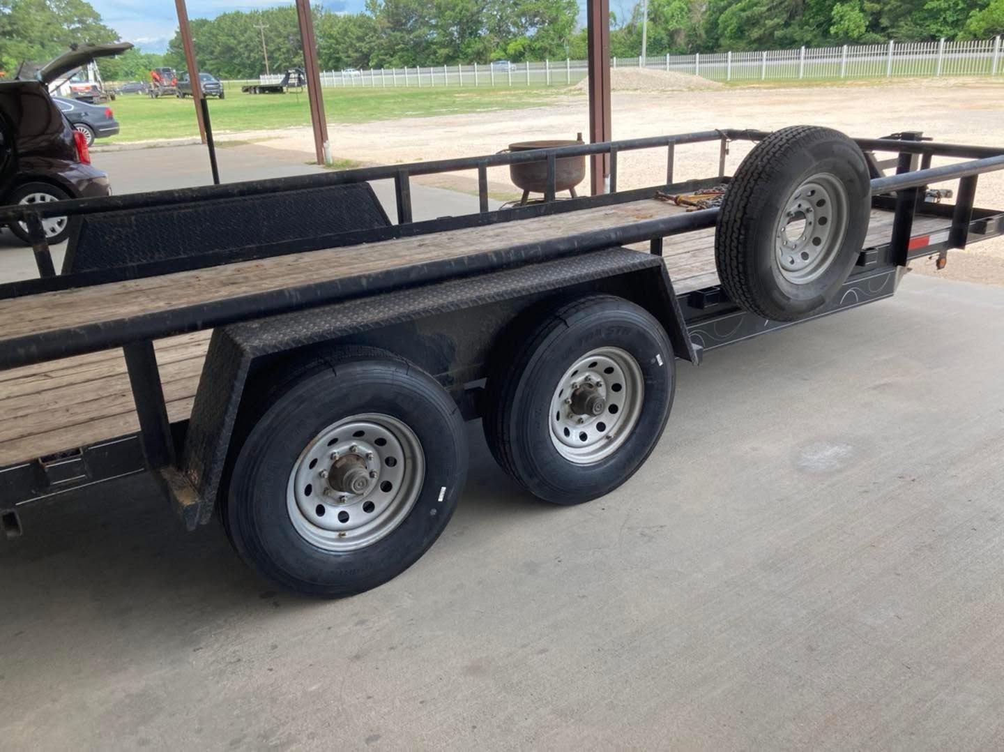 Black utility trailer with dual axles and spare tire parked under a pavilion outdoors