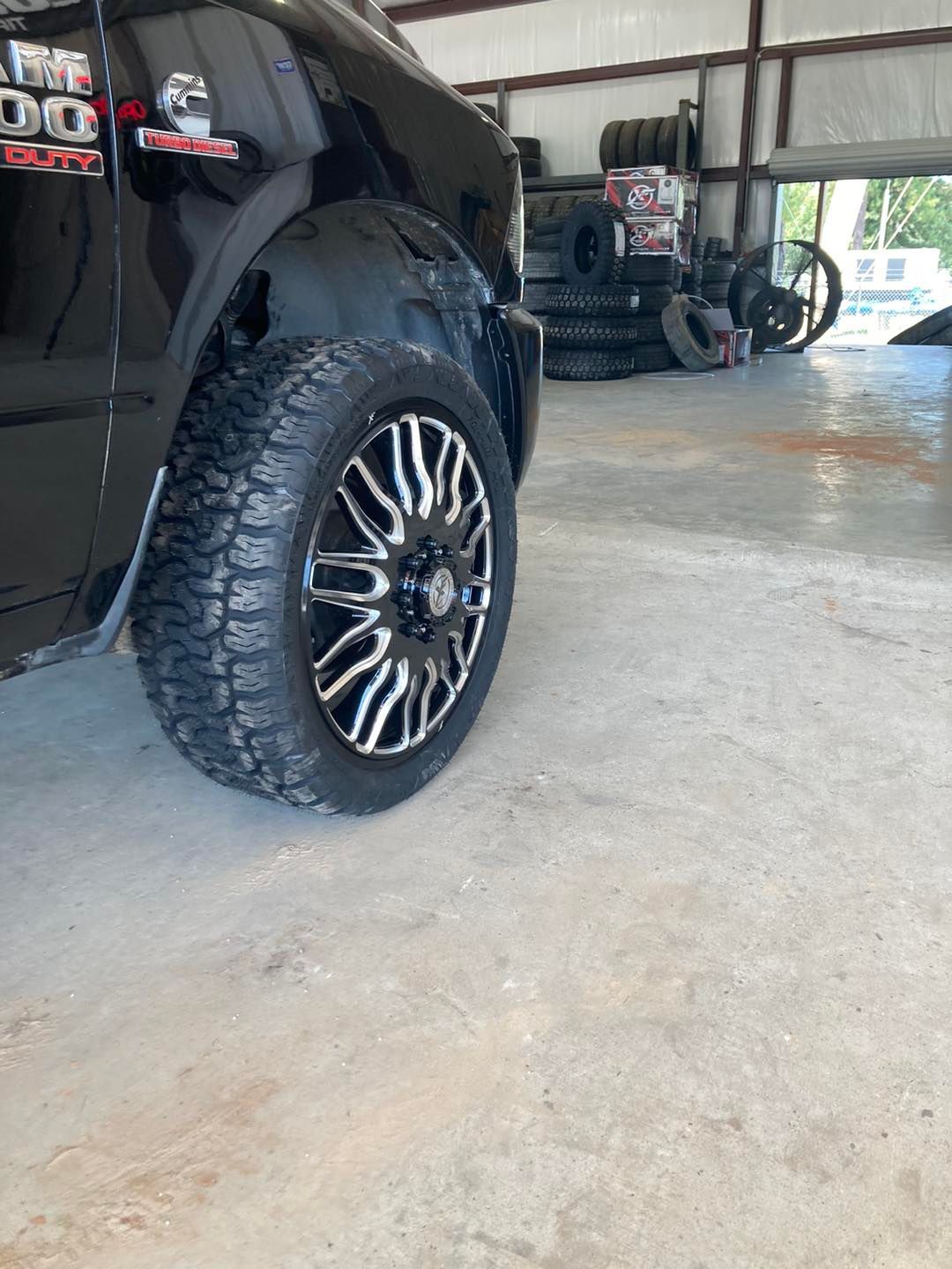 Black pickup truck with large chrome wheel in a garage, with another truck in the background