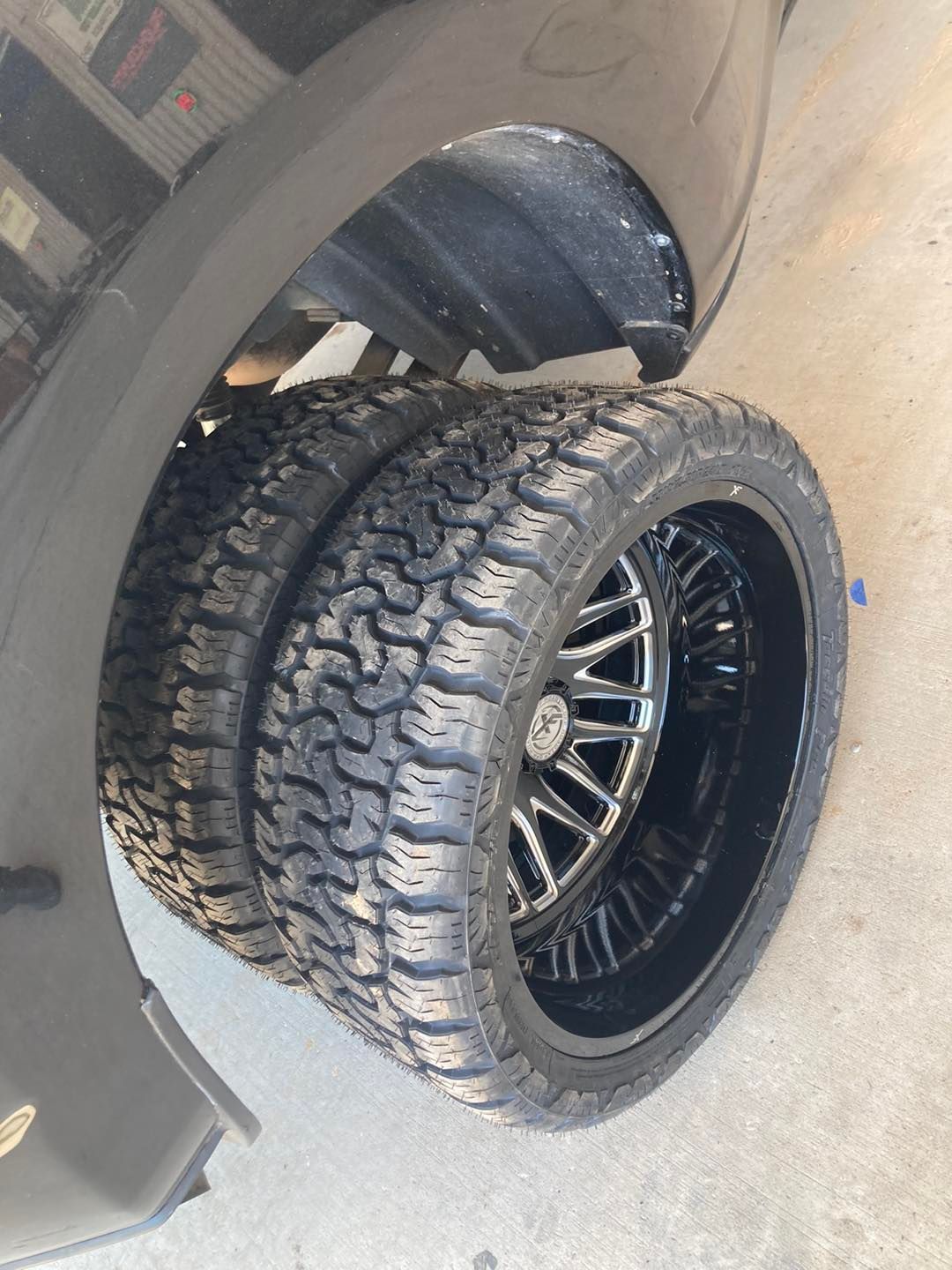 Piled off-road tires leaning against a black vehicle on a concrete driveway.