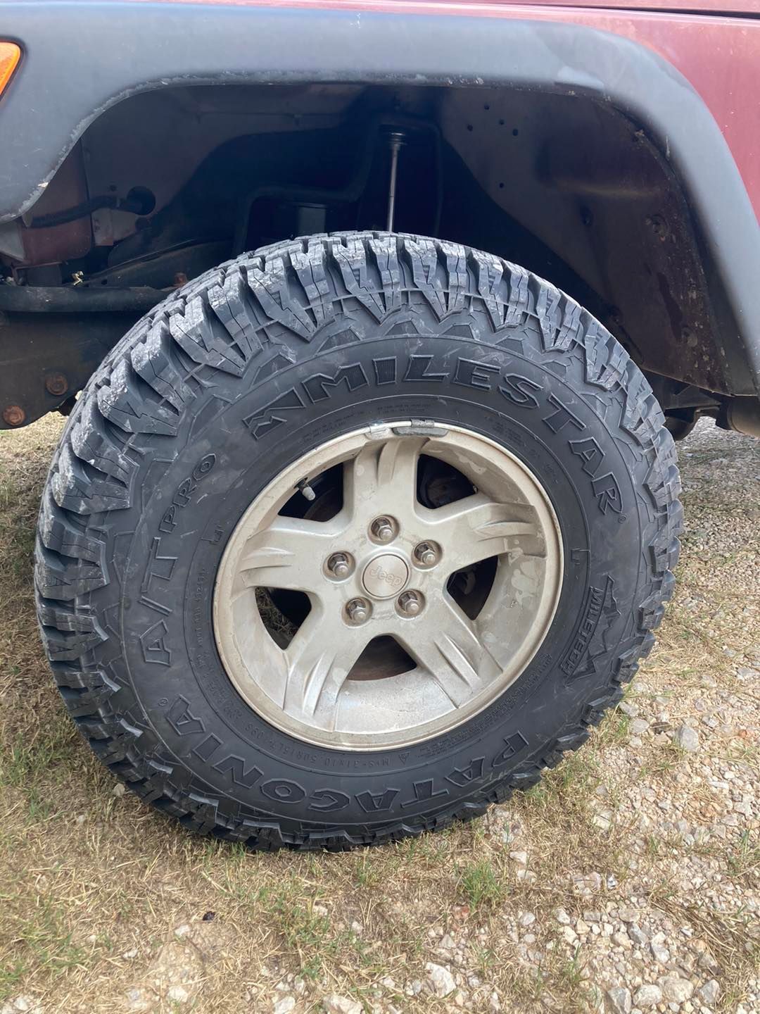 Off-road tire and silver rim on a vehicle, parked on gravel beside grass.