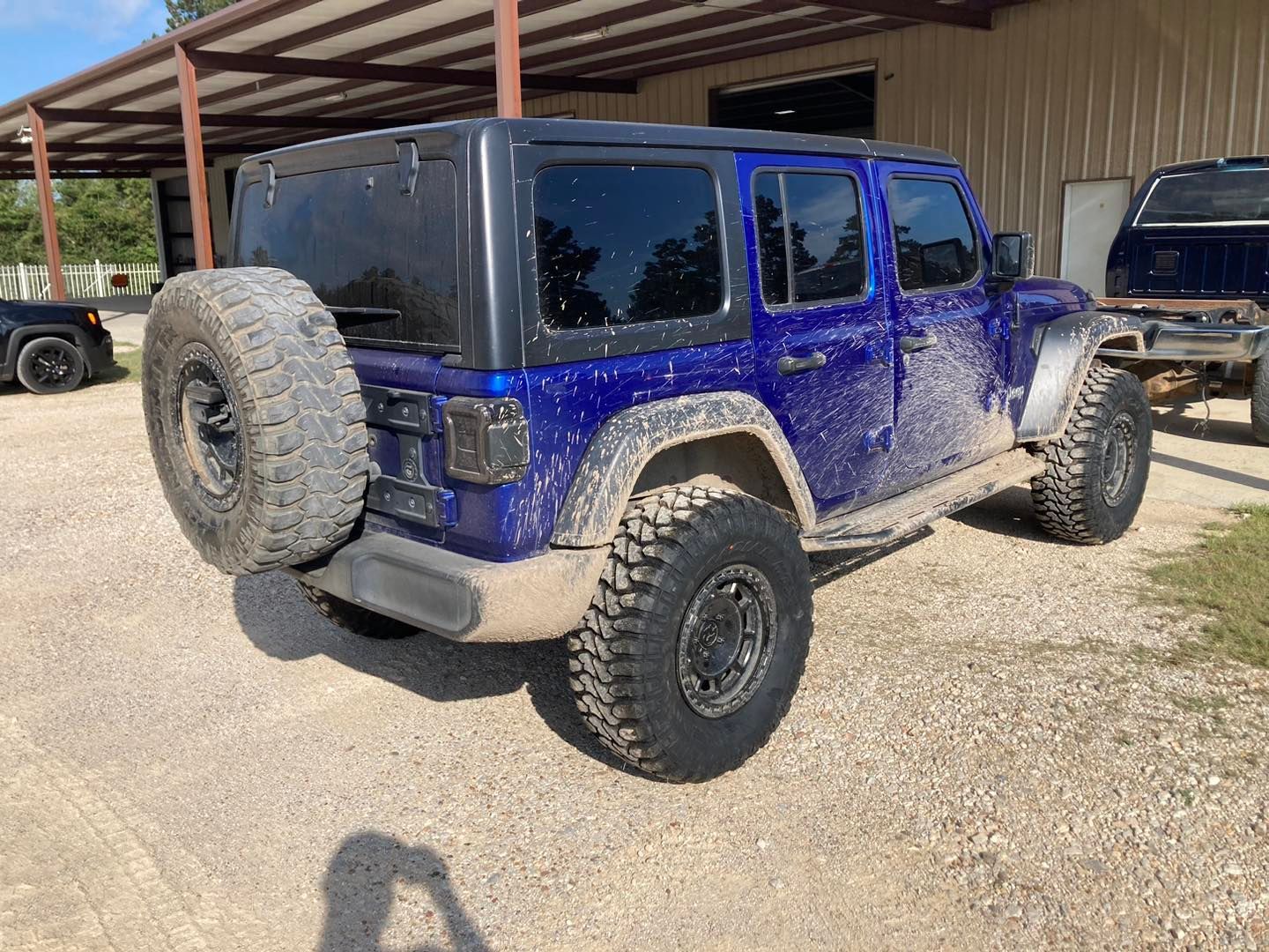 Blue Jeep Wrangler parked on gravel beside a metal shed, with a rear-mounted spare tire and off-road tires