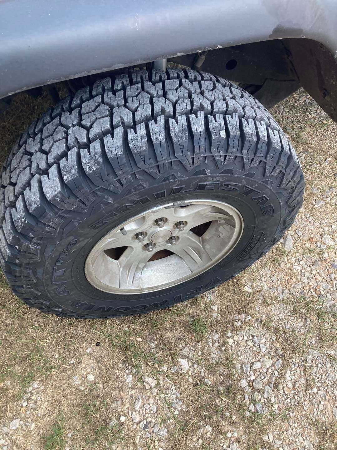 Close-up of a vehicle tire with an alloy rim on gravel, showing a rugged tread under the wheel well