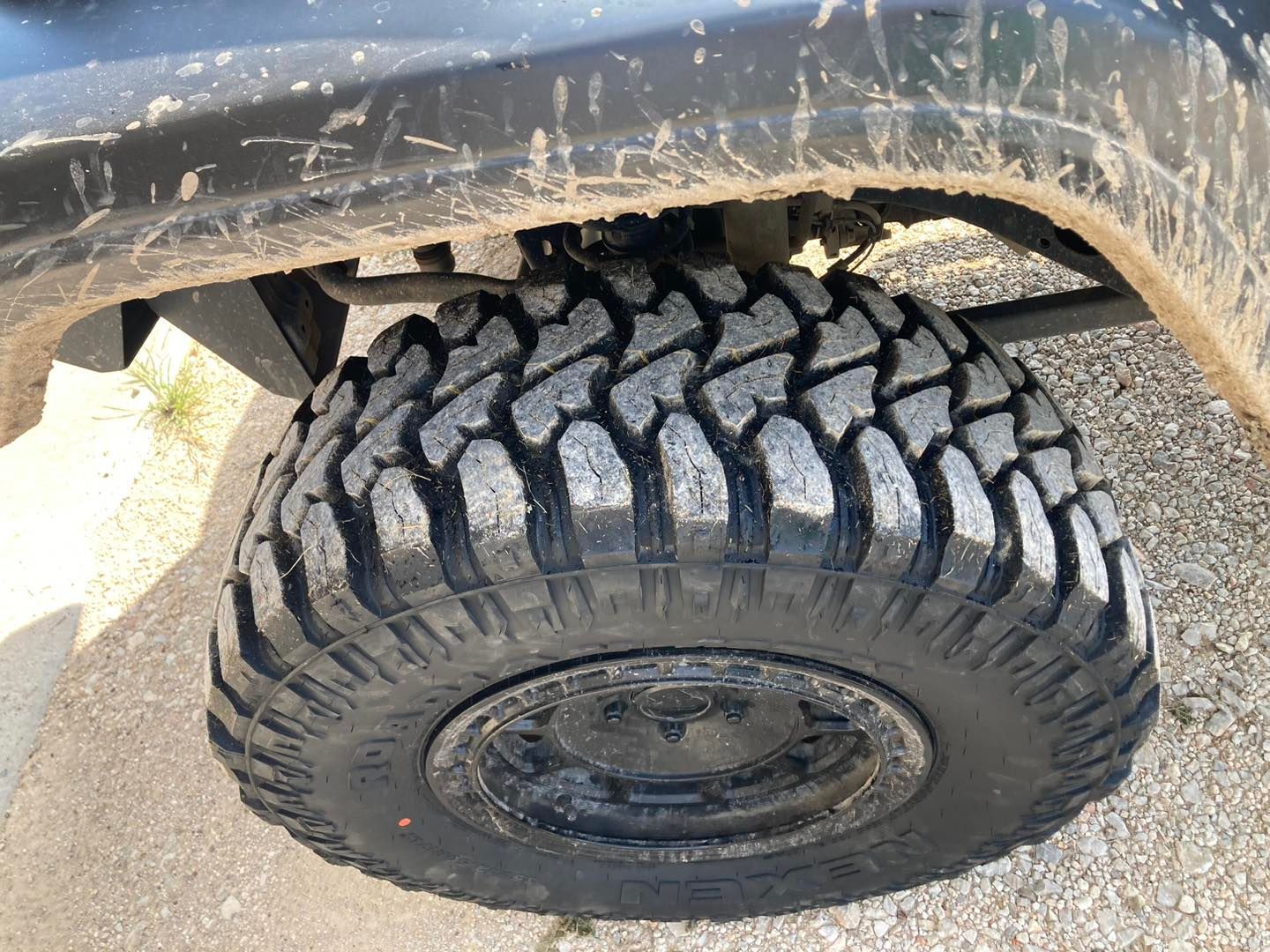 Mud-terrain tire under a vehicle’s wheel well on gravel, with a worn black fender above.