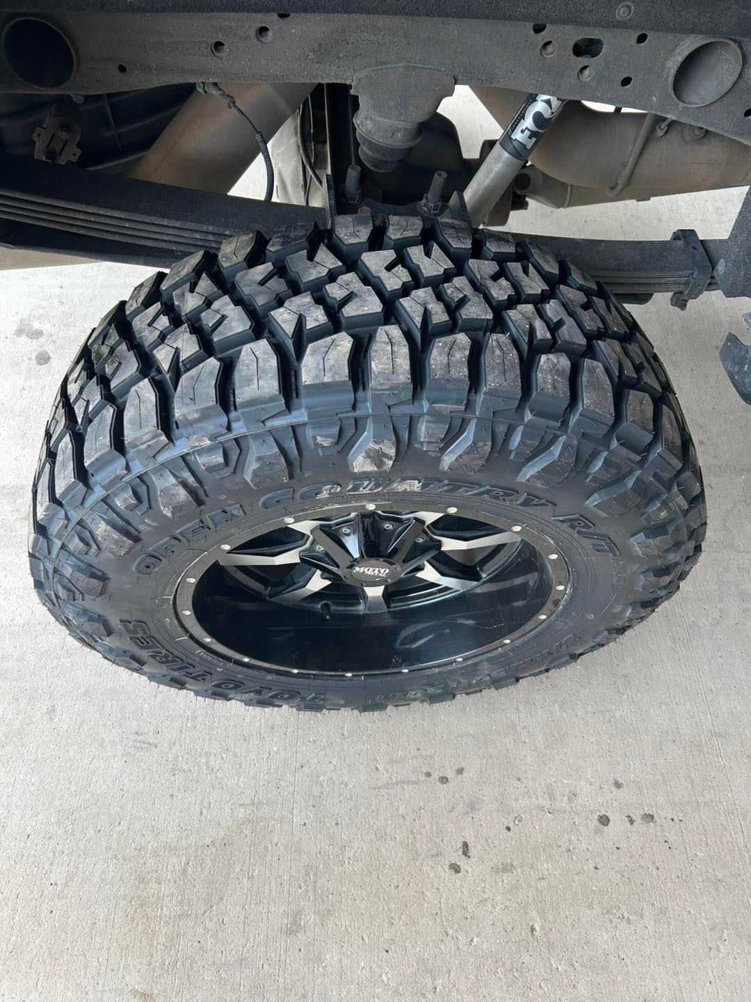 Off-road tire with black rim under a vehicle, viewed from the side on concrete.