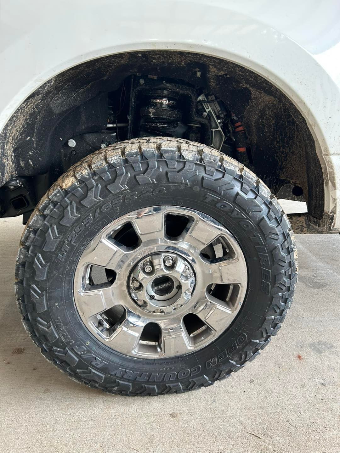 Close-up of a truck’s front wheel and tire on a garage floor, with white body and chrome rim