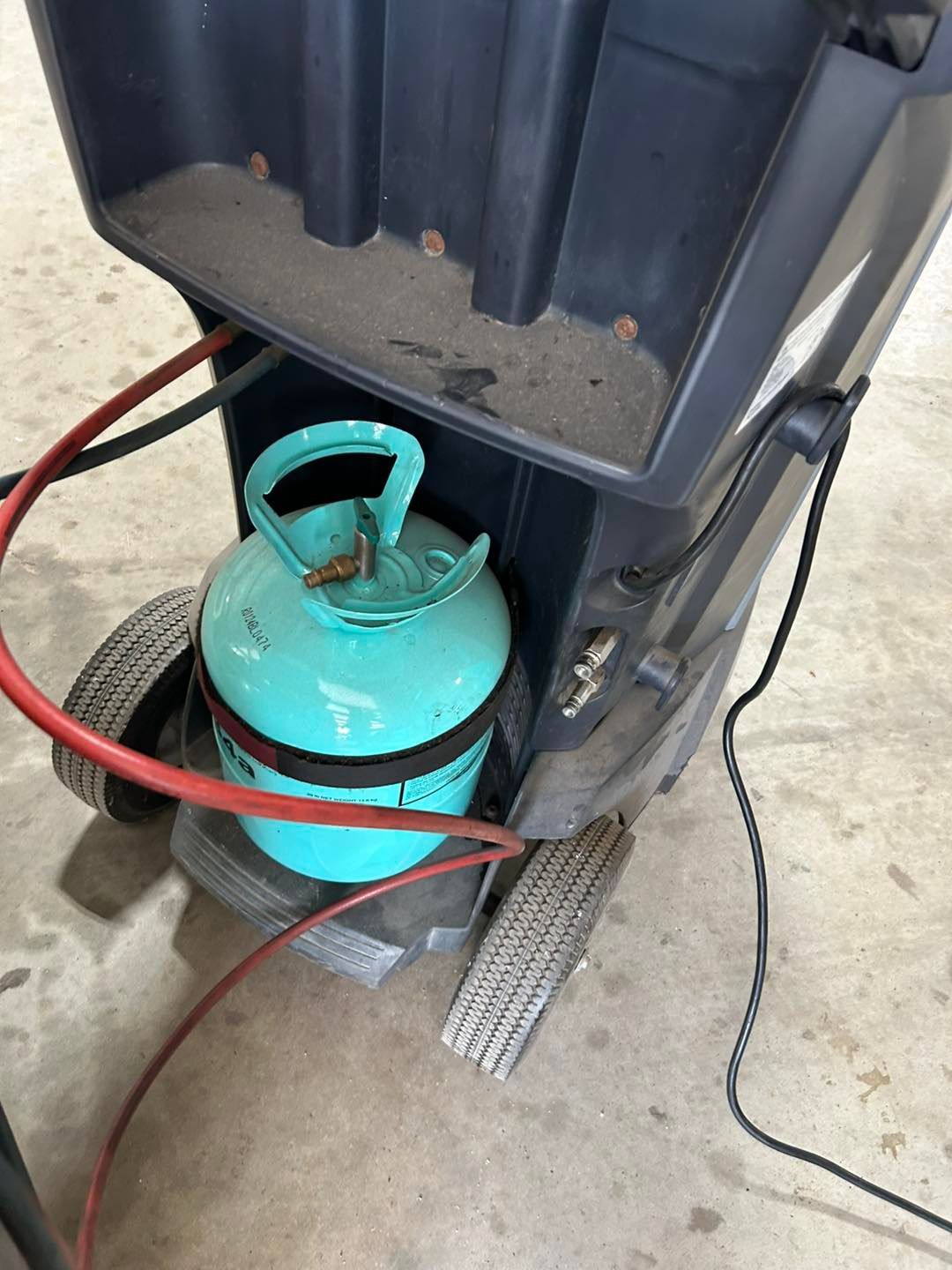 Teal gas cylinder strapped to a small cart under a dark machine in a workshop