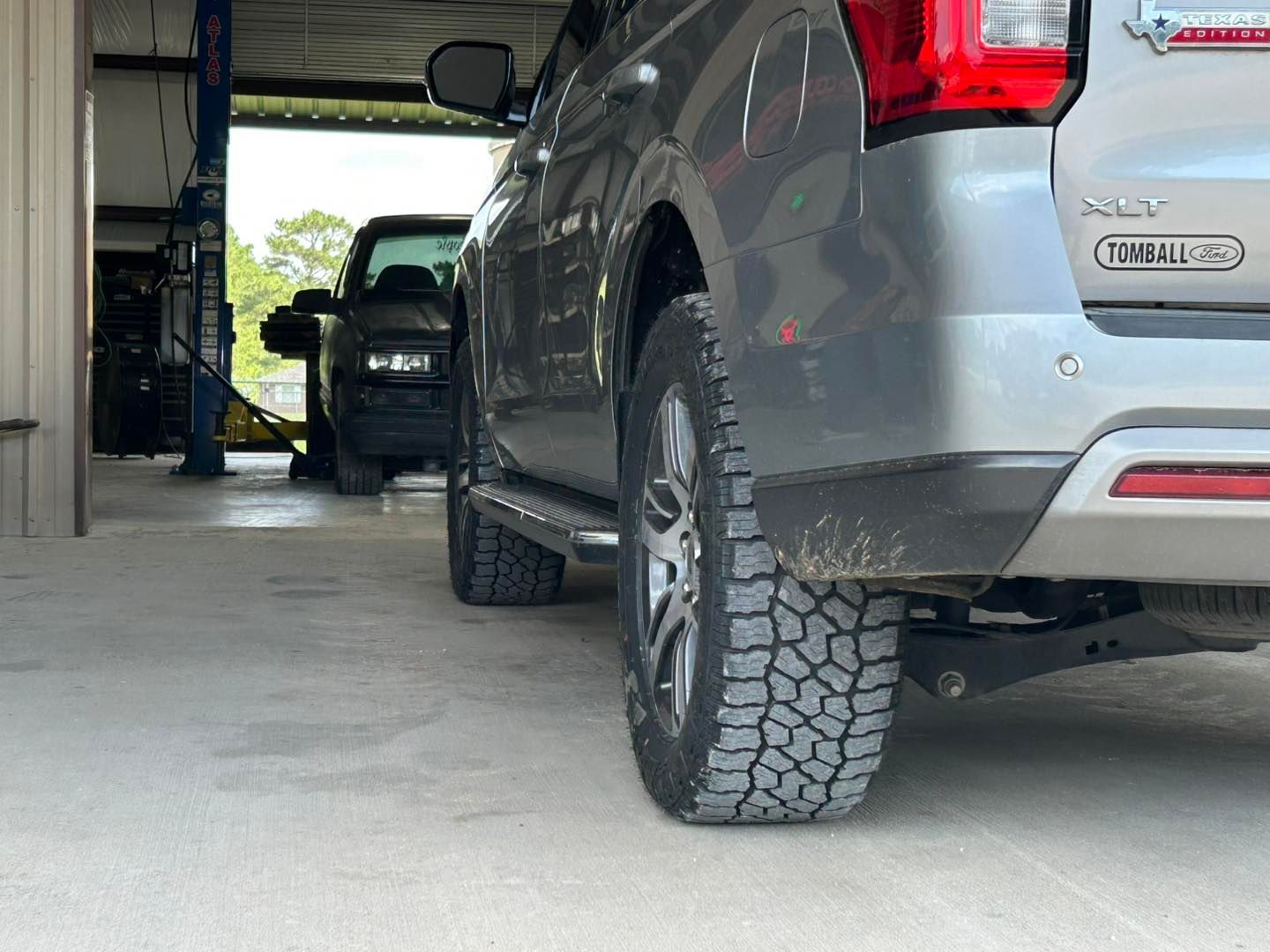 Gray SUV parked in a garage, rear view with off-road tires and a partially visible black vehicle inside.