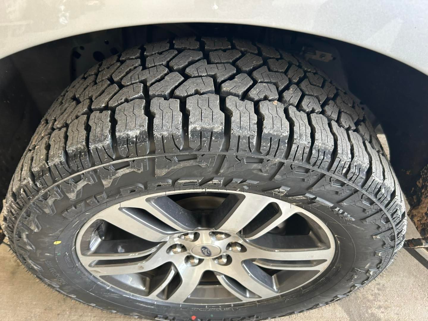 Car tire with a cracked, worn tread and silver alloy wheel under a white vehicle.