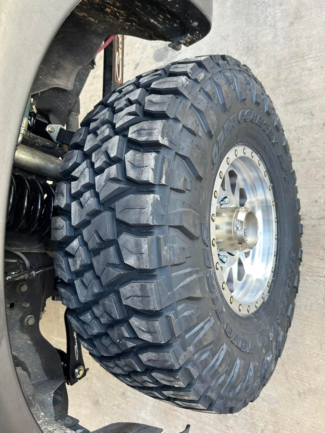 Off-road tire with deep tread on a polished wheel, shown close-up under a vehicle suspension.