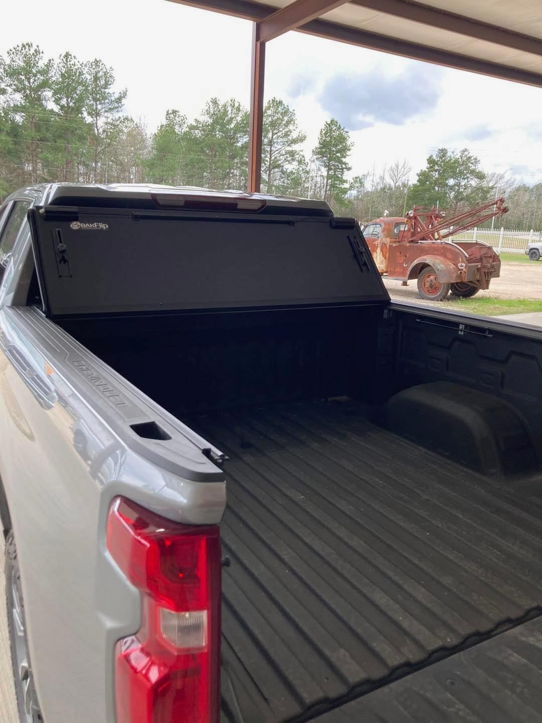 Black pickup truck bed with a hard tonneau cover, viewed from the rear under a carport.