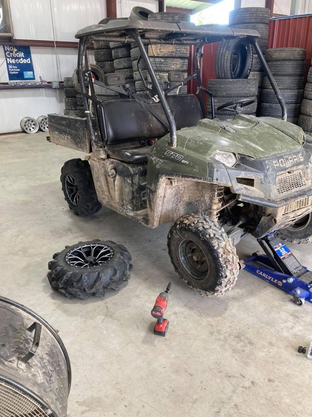 Mud-covered utility vehicle with a missing tire in a garage, with a jack and spare tire nearby